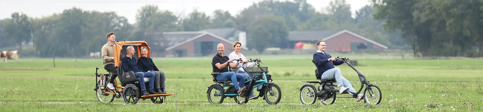 Drie aangepaste fietsen van vanRaam op een pad door een weiland: een riksja met twee passagiers, een zij-aan-zij-fiets en een driewieler.